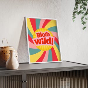 Könnte beinhalten: Ein gerahmter Kunstdruck mit weißem Rahmen. Das Kunstwerk zeigt ein lebendiges Design im Retro-Stil mit einem gelben Sonnenstrahl in der Mitte, umgeben von wirbelnden roten, rosa, türkisfarbenen und gelben Strahlen. Der Text "Bleib Wild!" ist in Rot und Weiß.