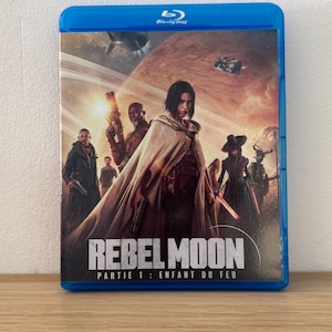 Peut inclure: Boîtier Blu-ray bleu pour "Rebel Moon: Partie 1 - Enfant du Feu". L'illustration de la couverture présente des personnages dans un cadre de science-fiction, avec le titre en évidence.