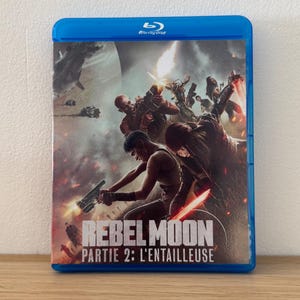 Peut inclure: Un boîtier Blu-ray bleu avec le film "Rebel Moon: Partie 2 : L'Entailleuse". La couverture présente une scène d'action de science-fiction avec des personnages armés et un fond enflammé. Le boîtier est posé sur une surface en bois.