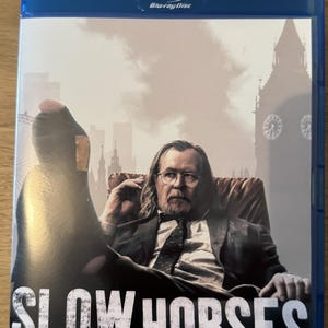 Peut inclure: Un boîtier Blu-ray bleu avec le titre "SLOW HORSES" en blanc. La couverture présente un homme en costume assis dans un fauteuil, avec un paysage urbain en arrière-plan. Le texte "SAISON CINQ" est également présent.