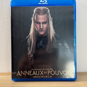 Peut inclure: Boîtier Blu-ray de "Le Seigneur des Anneaux : Les Anneaux de Pouvoir", Saison Deux. La couverture présente un portrait d'un personnage aux longs cheveux blonds et aux oreilles pointues. Le disque est dans un boîtier bleu.