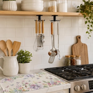 May include: A kitchen setting with a floral mat featuring butterflies, a gas hob, and kitchen tools. Wooden spoons and a jug are on the counter. Shelves display white bowls, jars, and potted plants. The colour scheme is soft and welcoming.