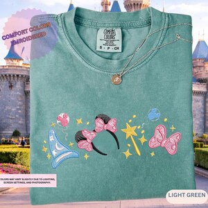 A green t-shirt with a variety of colorful and whimsical embroidered designs, including a wizard hat, butterflies, stars, and other decorative elements, displayed against a castle-like background.