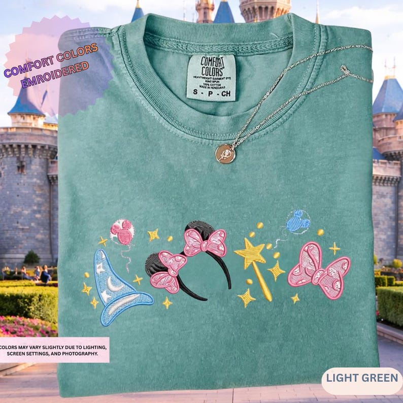 A green t-shirt with a variety of colorful and whimsical embroidered designs, including a wizard hat, butterflies, stars, and other decorative elements, displayed against a castle-like background.