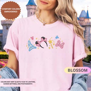 A woman wearing a pink t-shirt with a Disney-themed design, including a wizard hat, butterflies, and a flower, standing in front of a castle.