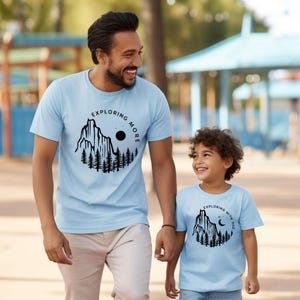 May include: Two light blue t-shirts with matching mountain and forest graphic designs. One shirt says "Exploring More" and the other says "Exploring with Dad". The shirts are worn by a man and a child.