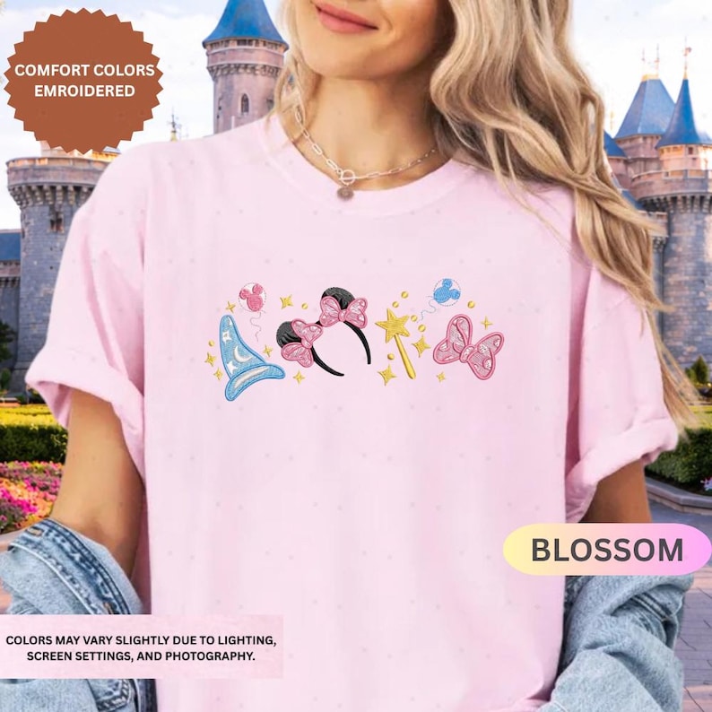 A woman wearing a pink t-shirt with a Disney-themed design, including a wizard hat, butterflies, and a flower, standing in front of a castle.