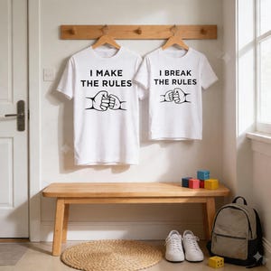 May include: Two white t-shirts with black text and a graphic of two fists bumping. One shirt says "I MAKE THE RULES", the other says "I BREAK THE RULES". The shirts hang on wooden hangers against a white wall.