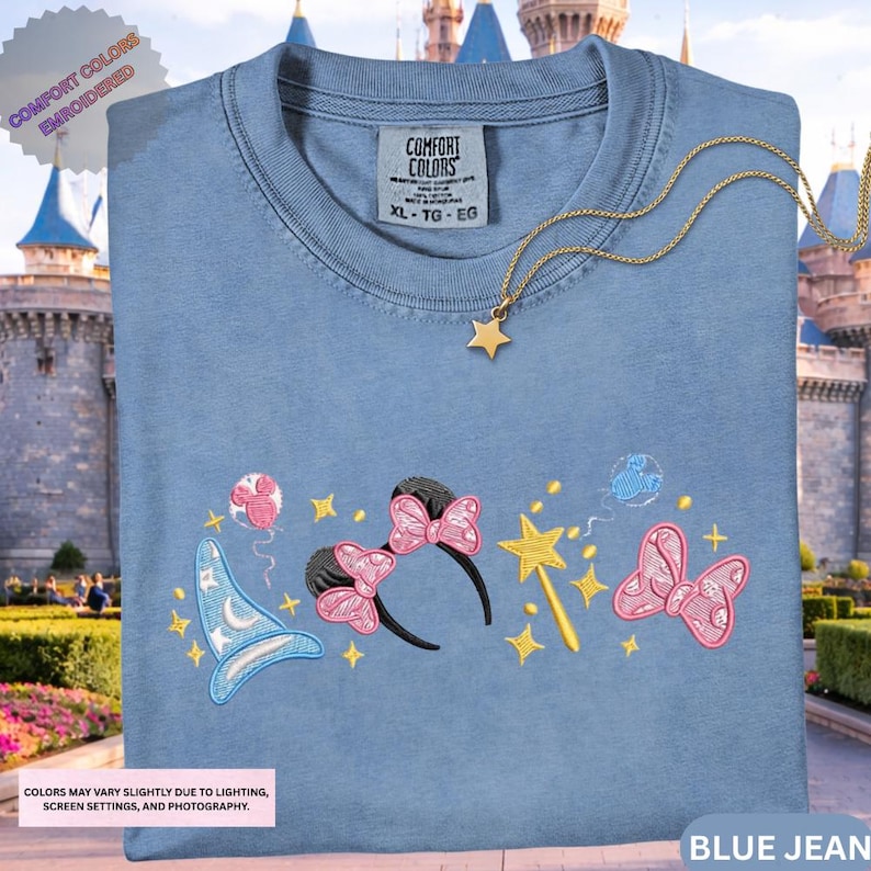 A blue t-shirt with various Disney-themed embroidered designs, including a wizard hat, butterflies, and stars, displayed in front of a castle.