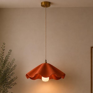 Könnte beinhalten: Eine orangefarbene Pendelleuchte mit gerüschtem Schirm hängt an einer goldfarbenen Deckenleuchte. Das Licht beleuchtet ein Wohnzimmer mit einem beigen Sofa, einem orangefarbenen Kissen und einem runden Couchtisch aus Holz. Ein gerahmtes abstraktes Kunstwerk hängt an der Wand.