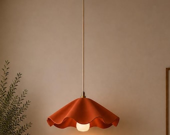 Puede incluir: Una lámpara colgante naranja con una pantalla con volantes cuelga de una lámpara de techo dorada. La luz ilumina una sala de estar con un sofá beige, un cojín naranja y una mesa de centro redonda de madera. Una obra de arte abstracta enmarcada cuelga de la pared.