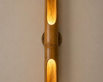 May include: A vertical wall sconce crafted from a single piece of natural bamboo, featuring two hollowed-out sections that emit a warm, golden light from the top and bottom. The bamboo has a smooth, light brown finish with visible wood grain.