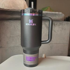 Puede incluir: Un vaso Stanley Quencher H2.0 FlowState negro con asa y pajita. El vaso tiene una tapa negra con borde plateado y el logotipo de Stanley. El texto "QUENCHER H TUMBLER" es visible en la etiqueta.
