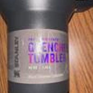 Puede incluir: Un vaso Stanley Quencher negro con asa. La etiqueta dice "Quencher Tumbler" en morado y azul. El vaso es de acero inoxidable y tiene una capacidad de 1,18 litros.