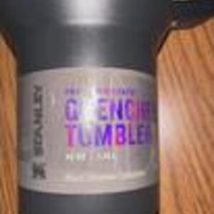 Puede incluir: Un vaso Stanley Quencher negro con asa. El vaso tiene una etiqueta que dice "Quencher Tumbler" en texto morado y azul. El vaso mide aproximadamente 24 cm de alto.