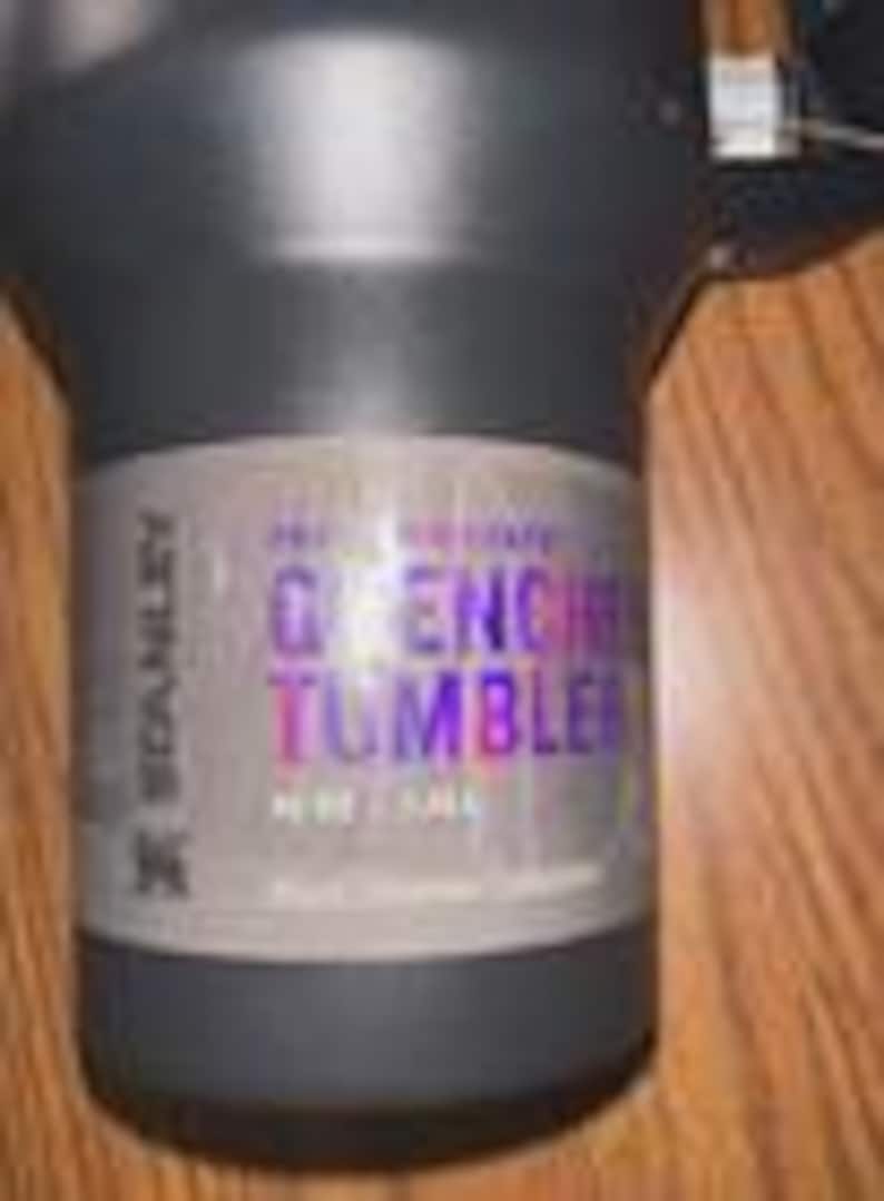 Puede incluir: Un vaso Stanley Quencher negro con asa. El vaso tiene una etiqueta que dice "Quencher Tumbler" en texto morado y azul. El vaso mide aproximadamente 24 cm de alto.