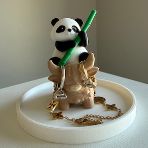 May include: A white and black panda figurine holding a green bamboo stalk, perched atop a tan-colored Highland cow figurine. The cow figurine is on a white tray, with gold-colored jewelry draped around it.
