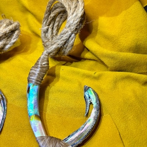 May include: A fish hook pendant crafted from iridescent abalone shell with a polished, multi-colored surface. The hook is wrapped with natural tan twine at the top and bottom, with a long, coiled length of the same twine attached to the top loop.