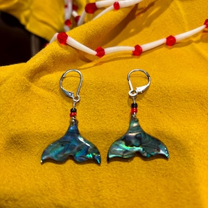 May include: A pair of silver earrings with iridescent blue-green abalone shell mermaid tail pendants. Each earring has a small cluster of red and black beads above the shell. The earrings are displayed on a yellow surface.