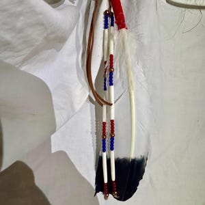 May include: A decorative hanging ornament featuring a large white feather with a black tip, accented by two vertical strands of white hair pipe beads interspersed with small red, blue, and metallic copper beads, tied with brown leather cords.