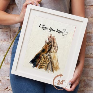 Puede incluir: Una obra de arte enmarcada con el texto "I Love You Mom" y tres ilustraciones de corazones. La obra representa una mano humana y dos patas de animales tocándose. El marco es blanco y la obra mide aproximadamente 61 cm.
