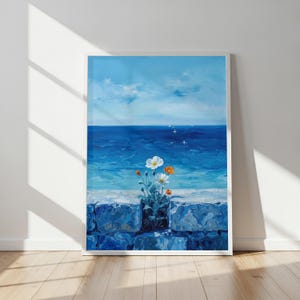 Puede incluir: Una impresión artística enmarcada que representa una escena oceánica con cielo azul y agua. Flores blancas y naranjas en una maceta oscura sobre un muro de piedra. La obra de arte está en un marco blanco.