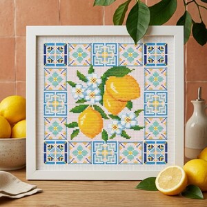 May include: A framed cross-stitch artwork featuring a lemon and floral design. The artwork showcases yellow lemons, white flowers, and green leaves against a pink background. The border has a blue and white tile pattern. The frame is white.