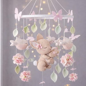 Puede incluir: Móvil para bebé con un conejito de peluche central que sostiene a un conejito más pequeño, rodeado de flores rosas, mariposas y hojas verdes. El móvil está suspendido de un anillo blanco con delicadas cuerdas, creando una decoración suave y caprichosa para la guardería.