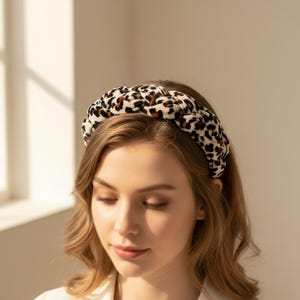 Könnte beinhalten: Ein dickes, geflochtenes Stirnband mit einer cremefarbenen Basis und einem braun-schwarzen Leopardenmuster. Das Accessoire hat eine weiche, plüschige Textur und eine breite, abgerundete Form, die auf dem Kopf getragen wird.