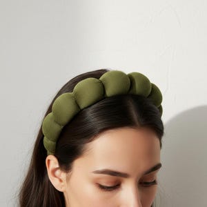 Könnte beinhalten: Olivgrünes, gepolstertes Stirnband mit einer Reihe von abgerundeten, stoffbezogenen Blasen. Das Stirnband wird auf dunklem Haar getragen, vor einem schlichten, weißen Hintergrund. Das Accessoire verleiht Textur und Farbe.