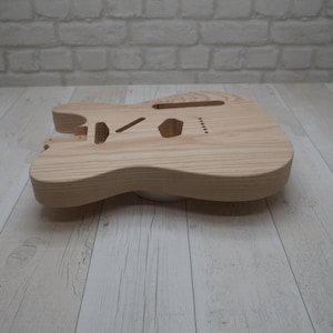 Pode incluir: Um corpo de guitarra elétrica de madeira clara sem acabamento com um padrão de grão visível. O corpo apresenta cavidades pré-cortadas para a ponte, captadores e eletrônicos, juntamente com um encaixe para o braço e orifícios para cordas.