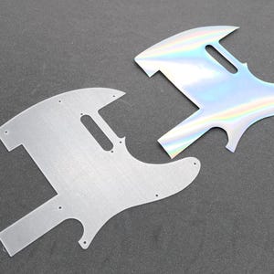 Könnte beinhalten: Zwei E-Gitarren-Schlagbretter in Telecaster-Form. Ein Schlagbrett hat eine gebürstete silberne Metalloberfläche mit sichtbaren Schraublöchern, während das andere eine reflektierende, schillernde holografische Oberfläche aufweist, die ihre Farbe verändert.