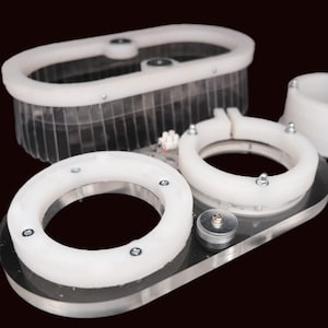 Könnte beinhalten: Eine Ansammlung weißer und klarer Kunststoffkomponenten, darunter kreisförmige und ovale Formen, mit Metallbefestigungen. Die Teile sind auf einer transparenten Acrylbasis vor dunklem Hintergrund angeordnet. Die Komponenten scheinen Teil eines größeren Mechanismus zu sein.