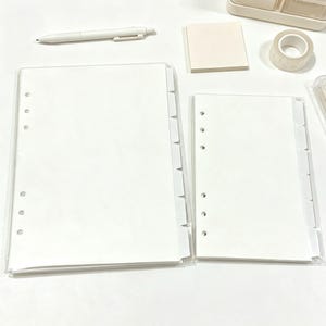 May include: Two sets of white, multi-hole punched planner inserts with side tabs, accompanied by a white retractable pen, a square stack of light beige sticky notes, and a roll of clear adhesive tape, all arranged for organized stationery use.