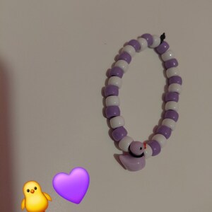Op de afbeelding: Een kralenarmband met afwisselend witte en lavendelkleurige kralen. Een lavendelkleurige eendenbedel met een zwarte kraag en oranje snavel hangt aan de armband. Een gele kuiken- en paarse hart-emoji zijn ook aanwezig.