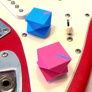May include: Close-up of a red and white electric guitar with two geometric knobs. One knob is blue with a pink stripe, and the other is pink with a blue stripe. The guitar has white pickups and a white switch.