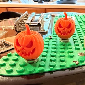 May include: A black electric guitar featuring a custom green building block style pickguard. Two orange 3D-printed jack-o'-lantern pumpkin knobs are attached to the guitar controls, replacing standard knobs, alongside a white plastic switch tip.