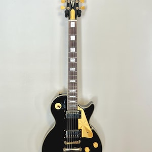Puede incluir: Una guitarra eléctrica negra con herrajes dorados, incluyendo clavijas, perillas y un golpeador. La guitarra tiene un acabado brillante y un diseño clásico, con el nombre de la marca en la pala.
