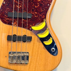 May include: A natural wood finish electric bass guitar featuring a tortoiseshell pickguard, two black EMG pickups, and a chrome bridge. The control plate is decorated with three bright yellow banana-shaped knobs arranged in a vertical row.