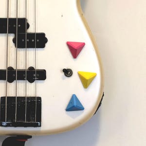 May include: Close-up of a white electric bass guitar with black pickups and strings. Three colorful, triangular knobs in pink, yellow, and blue are affixed to the body of the instrument.