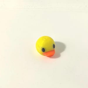 Puede incluir: Una pequeña figura tridimensional de cabeza de pato amarilla que presenta un pico naranja brillante y dos ojos circulares de color negro sólido. El objeto tiene una forma suave y redondeada, presentada como una pieza de cabeza aislada.