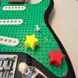 Op de afbeelding: Een zwarte elektrische gitaar met een groene, getextureerde slagplaat. De slagplaat heeft een Lego-achtig ontwerp en heeft een gele en een rode ster. De body van de gitaar is zwart en de hals is van hout.