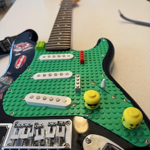 Puede incluir: Una guitarra eléctrica negra con un cuerpo verde cubierto de Lego. La guitarra presenta pastillas blancas, perillas de cabeza Lego amarillas y un puente plateado. El mástil es de madera y la correa de la guitarra es negra.