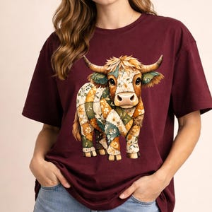 May include: Burgundy t-shirt with a Highland cow design. The cow is decorated with a patchwork of floral and geometric patterns in orange, green, and white. The shirt has short sleeves. The design is a colourful, eye-catching image.