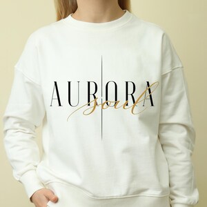 May include: White sweatshirt featuring the text "AURORA Soul" in black and gold script. The word "AURORA" is in a bold, sans-serif font, with a vertical line above and below. "Soul" is in a cursive font.