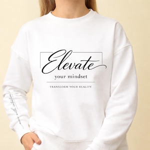 Op de afbeelding: Witte sweatshirt met het woord "Elevate" in zwart script, met daaronder de zin "your mindset". De zin "Transform your reality" is eronder gedrukt. De mouw heeft de woorden "move with intention".