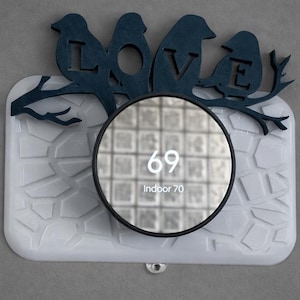 Puede incluir: Un dispositivo doméstico inteligente rectangular blanco con una pantalla circular negra que muestra los números "69" e "Indoor 70". Encima de la pantalla hay un recorte de madera azul oscuro de pájaros posados en una rama con la palabra "LOVE".