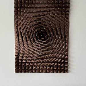 May include: Brown geometric wall art with a spiral design. The artwork features a series of triangular shapes arranged in a swirling pattern, creating a three-dimensional effect. The piece is mounted on a white wall.
