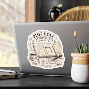 May include: A die-cut sticker featuring an open book with a hole in the page, a cup of coffee, a fountain pen, and sticky notes. The text reads PLOT HOLE APPRECIATION SOCIETY, we'll fix it in revisions, PLOT MAP, and MEETINGS ADJOURNED.