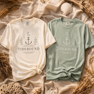 May include: Two short-sleeve crew neck t-shirts, one cream and one sage green. Each shirt features a central graphic with a crescent moon, an anchor, two leafy branches, and wavy lines above the text TIDEBOUND printed in a dark color.